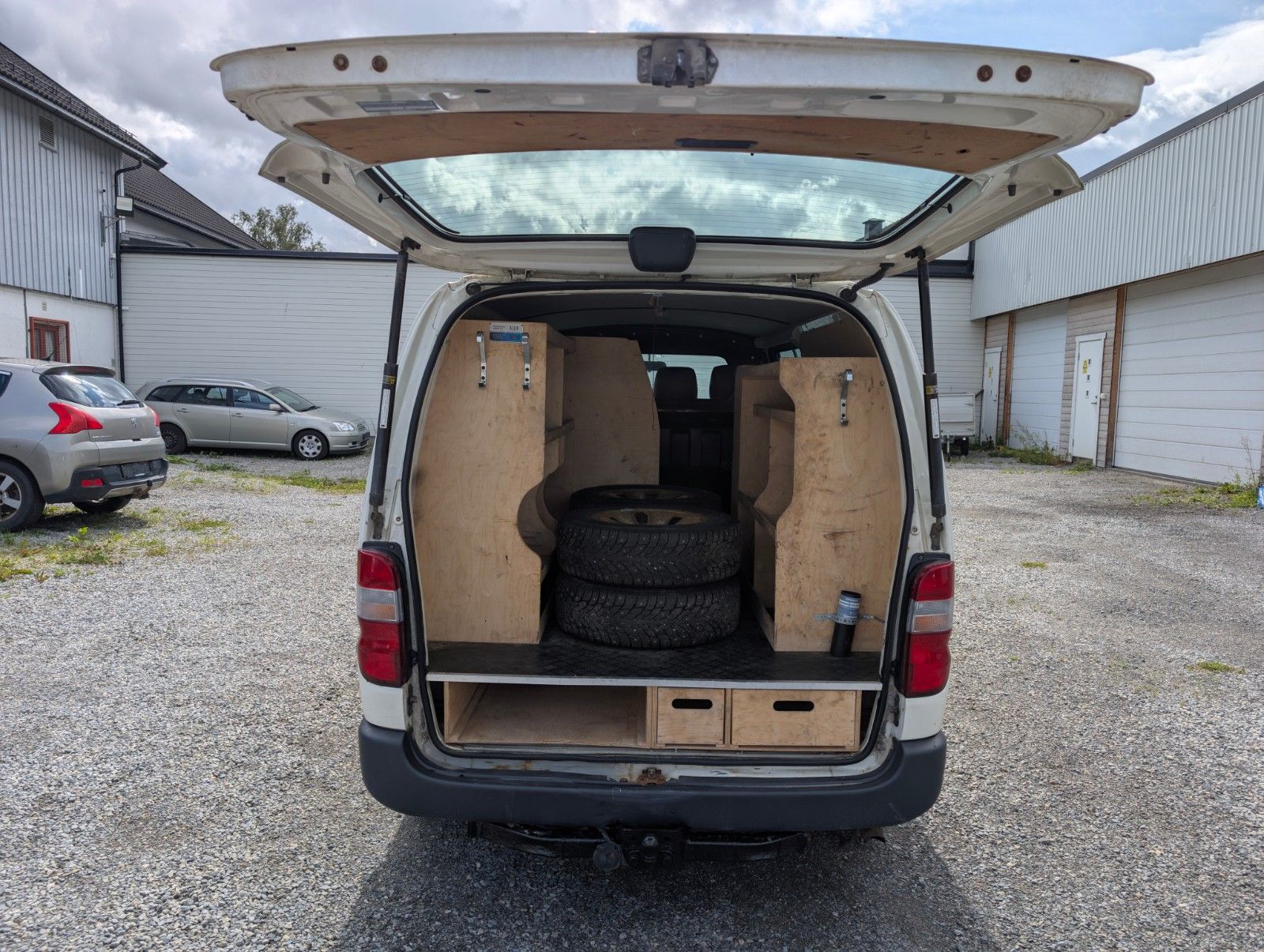 Toyota HiAce 4WD 2011 med åpen bakluke, treinnredning og ekstra dekk i bagasjerommet.