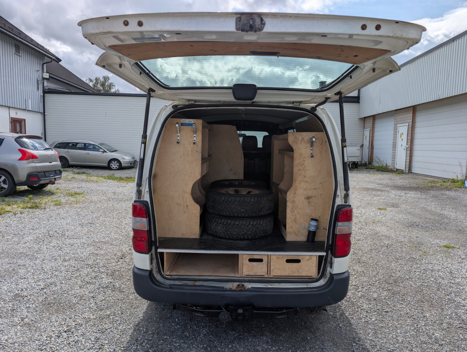 Toyota HiAce 4WD 2011 med åpen bakluke, treinnredning og ekstra dekk i bagasjerommet.